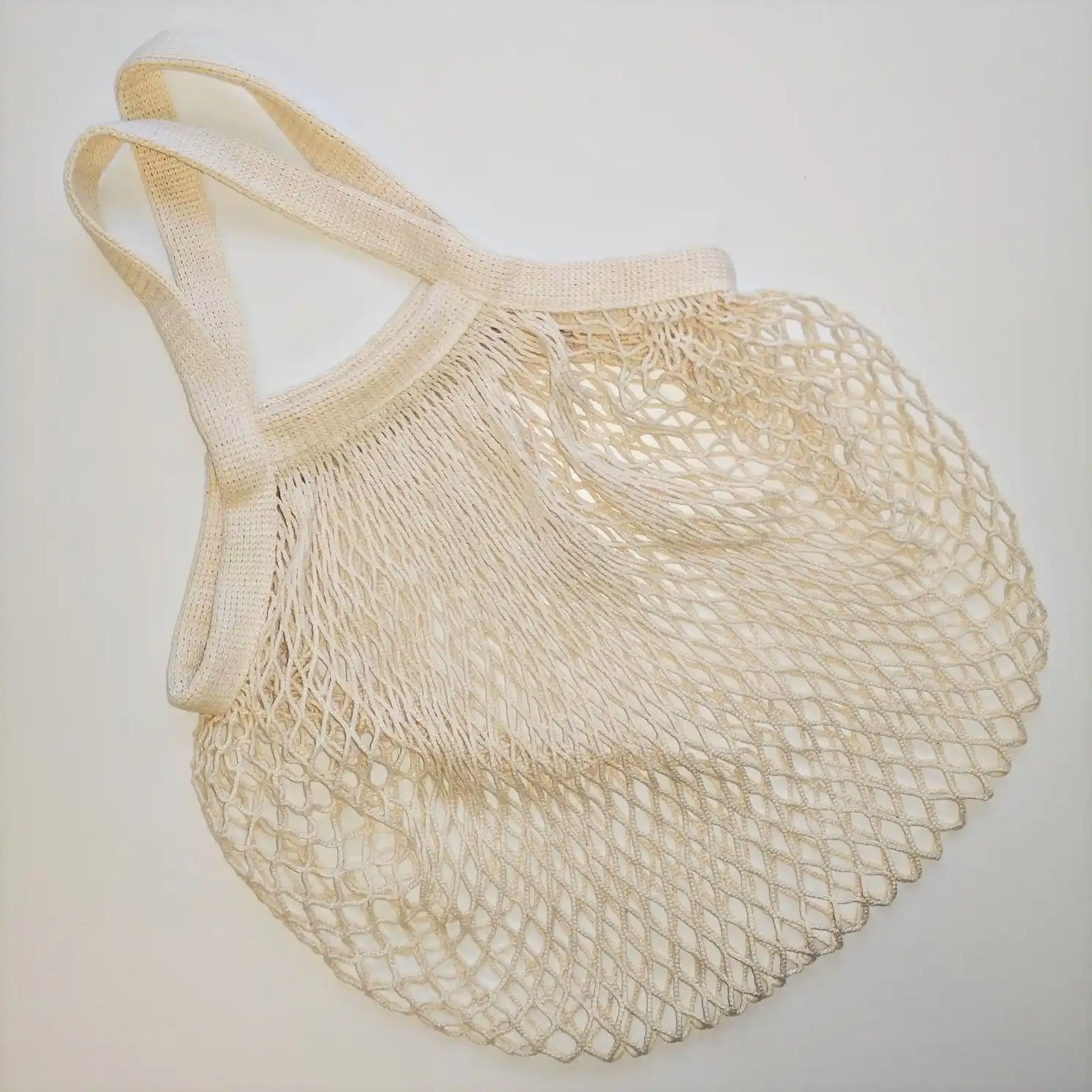 Beiger, wiederverwendbarer Netzbeutel mit zwei Henkeln mit mehreren Zitronen und einer Orange liegt auf einem Betonboden vor einer weißen Wand.