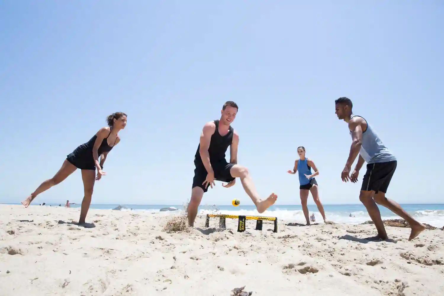 Vier Personen spielen Spikeball am Strand, gelber Ball über dem Netz, sonniger Himmel und Meer im Hintergrund