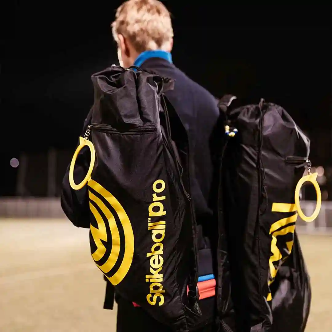 Person trägt zwei schwarze Spikeball Pro Rucksäcke mit gelbem Logo auf einem beleuchteten Sportplatz bei Nacht
