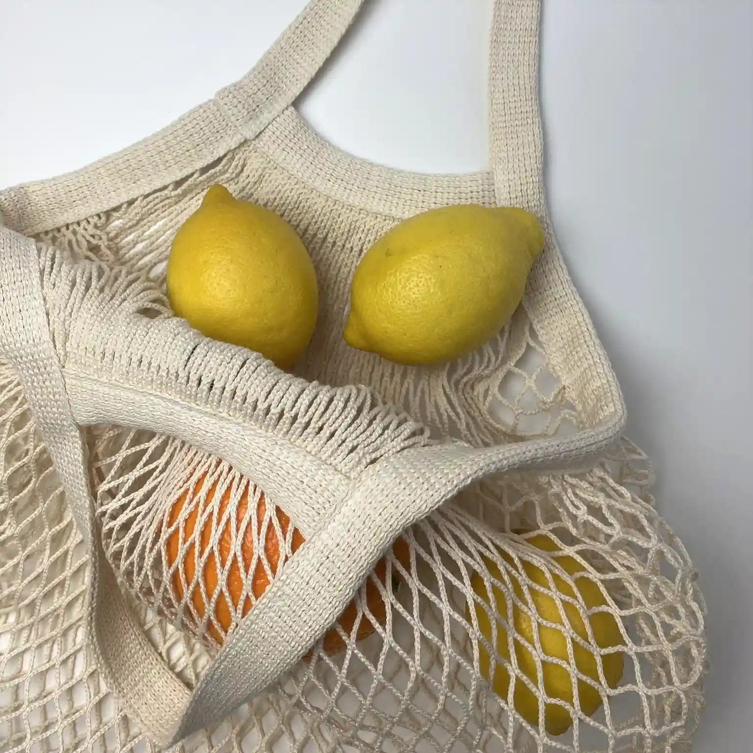 Beiger, wiederverwendbarer Netzbeutel mit zwei Henkeln liegt auf einem Betonboden vor einer weißen Wand.