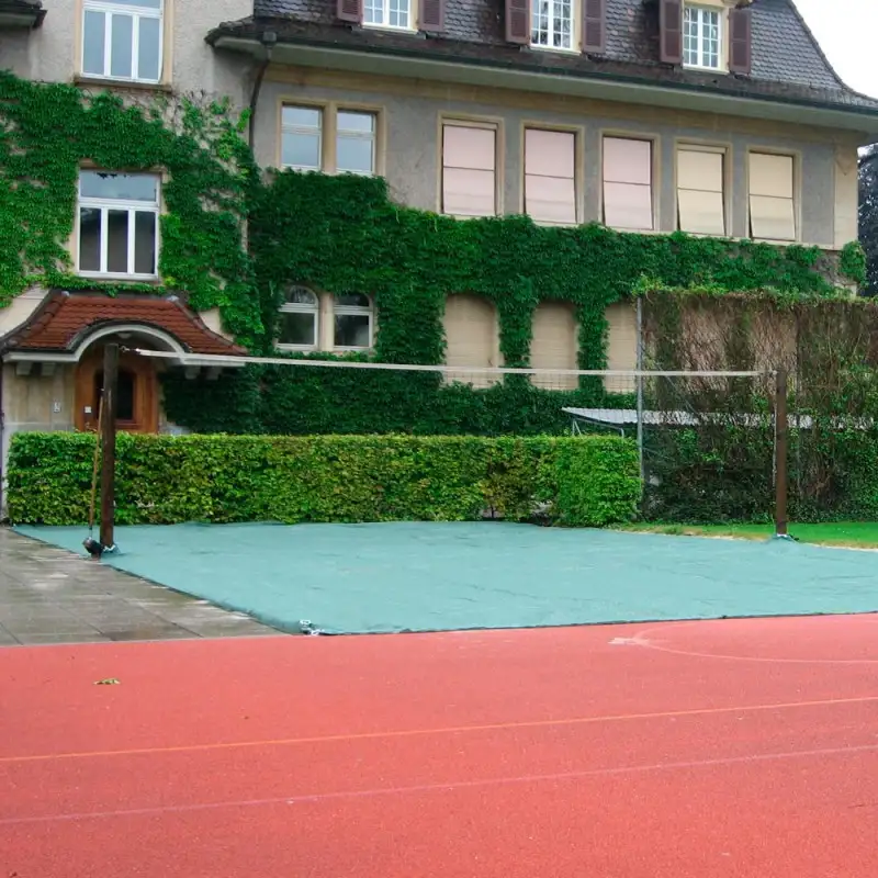 Außenansicht eines mit Efeu bewachsenen Gebäudes mit mehreren Fenstern. Davor steht ein Volleyballnetz; der Sandbereich ist mit einem grünen Polyethylengewebe abgedeckt. Im Vordergrund verläuft eine rote Laufbahn.
