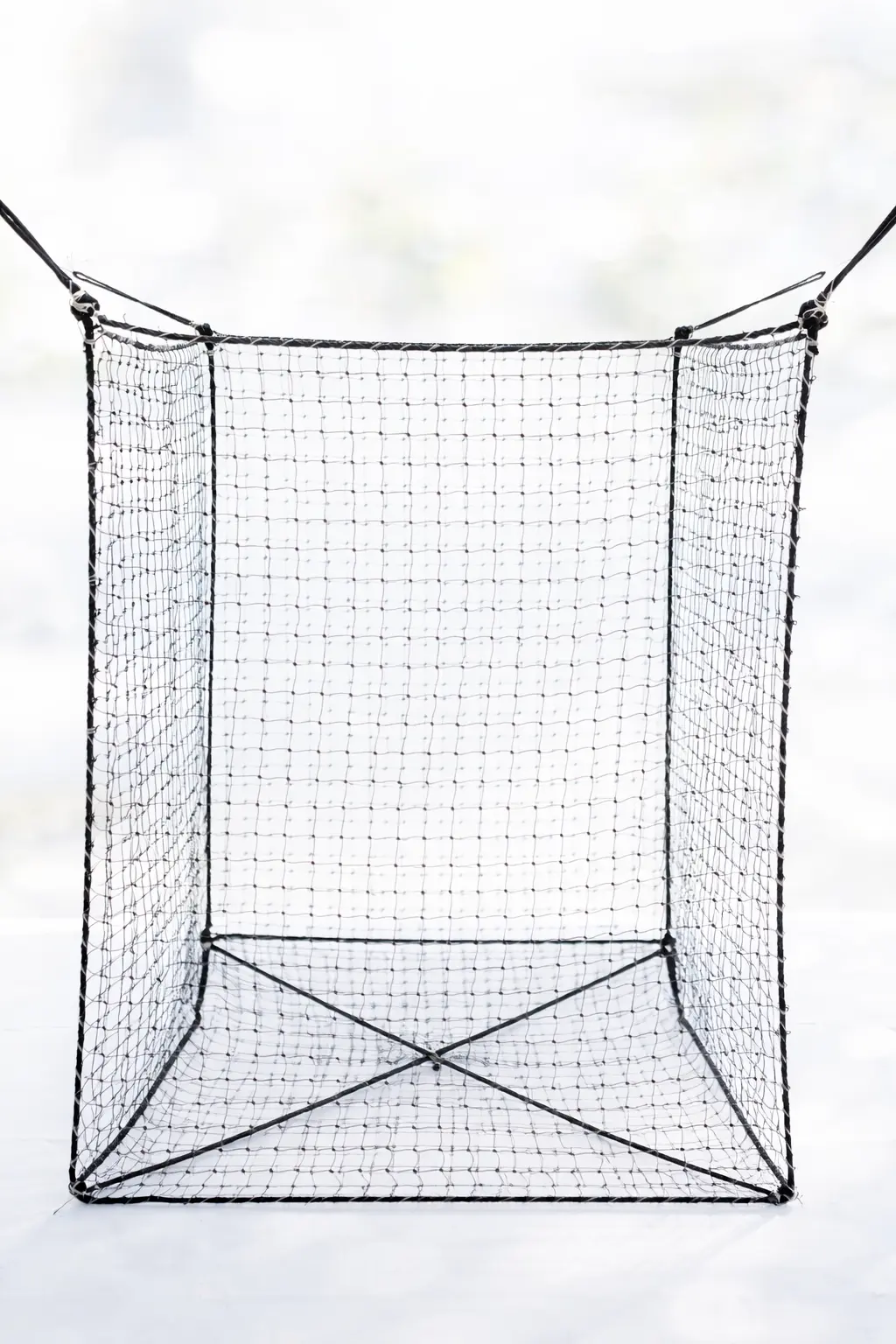 Großer würfelförmiger Netzkorb Woodbag aus schwarzen Schnüren steht auf einem Betonboden vor einer weißen Wand. Die Unterseite ist mit überkreuzten Verstärkungsseilen gesichert