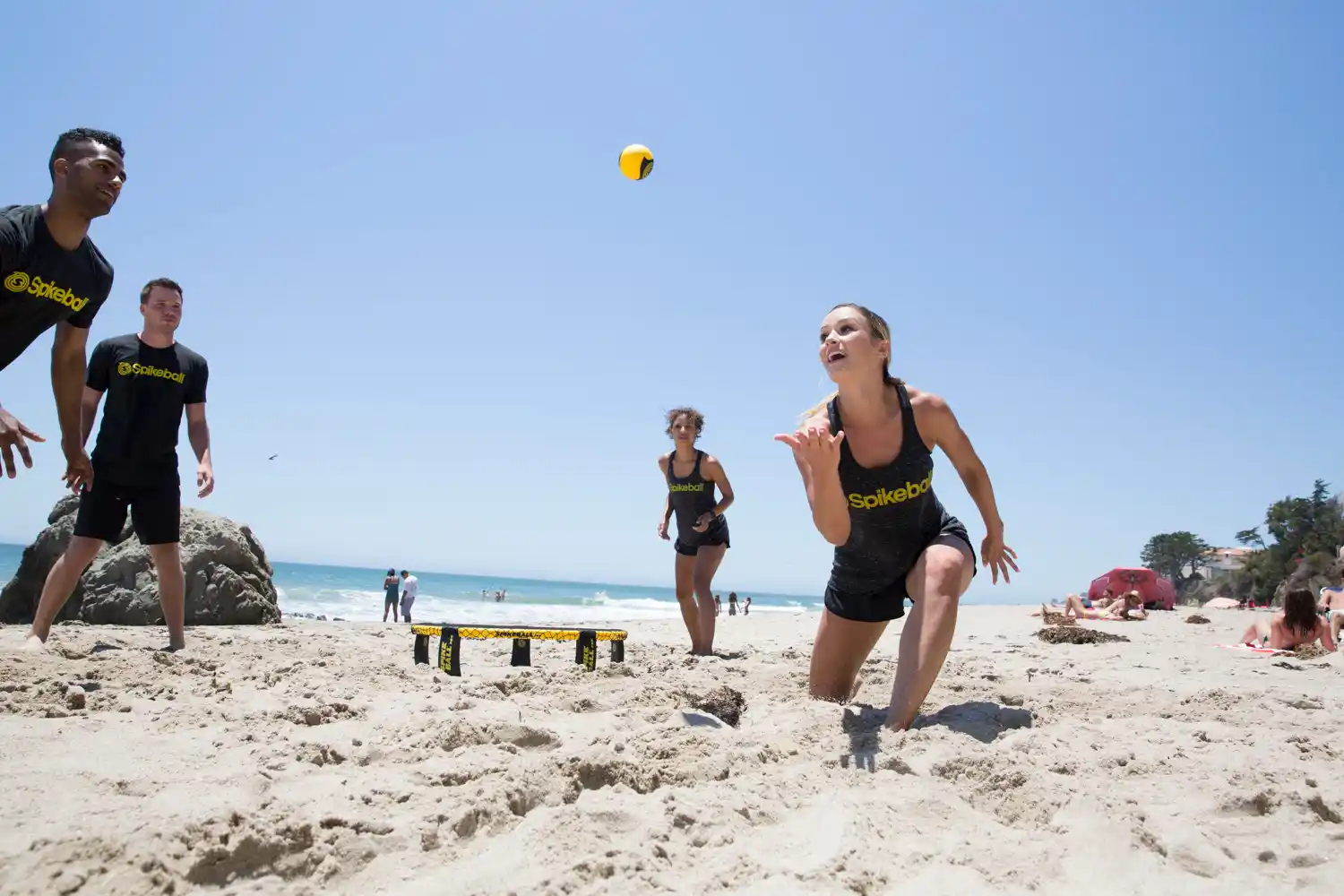 Vier Personen spielen Spikeball am Strand, gelb‑schwarzer Ball über dem Netz, sonniger Himmel