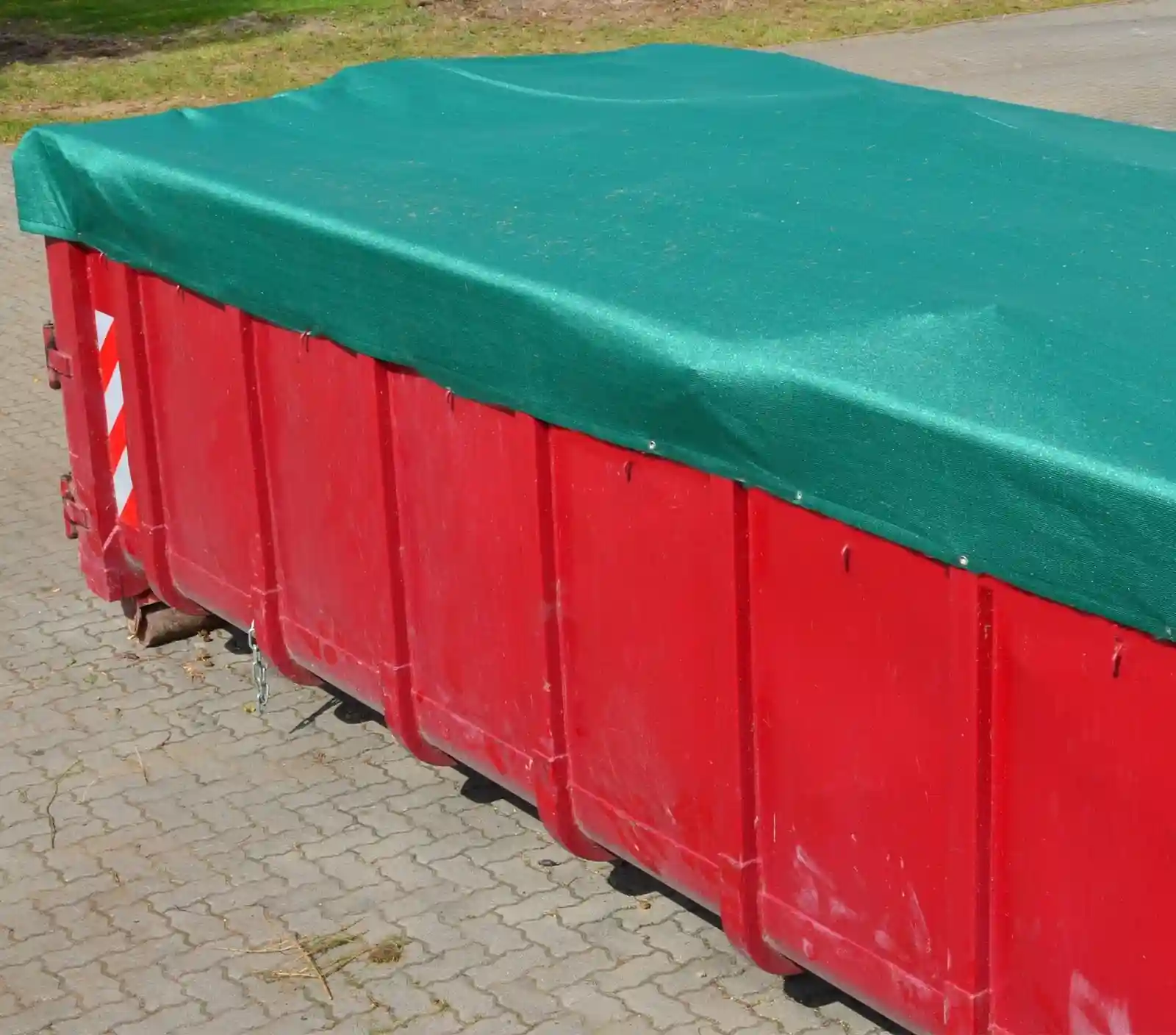 Großer roter Container mit einer straff gespannten grünen Abdeckplane, aufgestellt auf einem gepflasterten Untergrund.  Im Hintergrund ist eine Grünfläche zu sehen