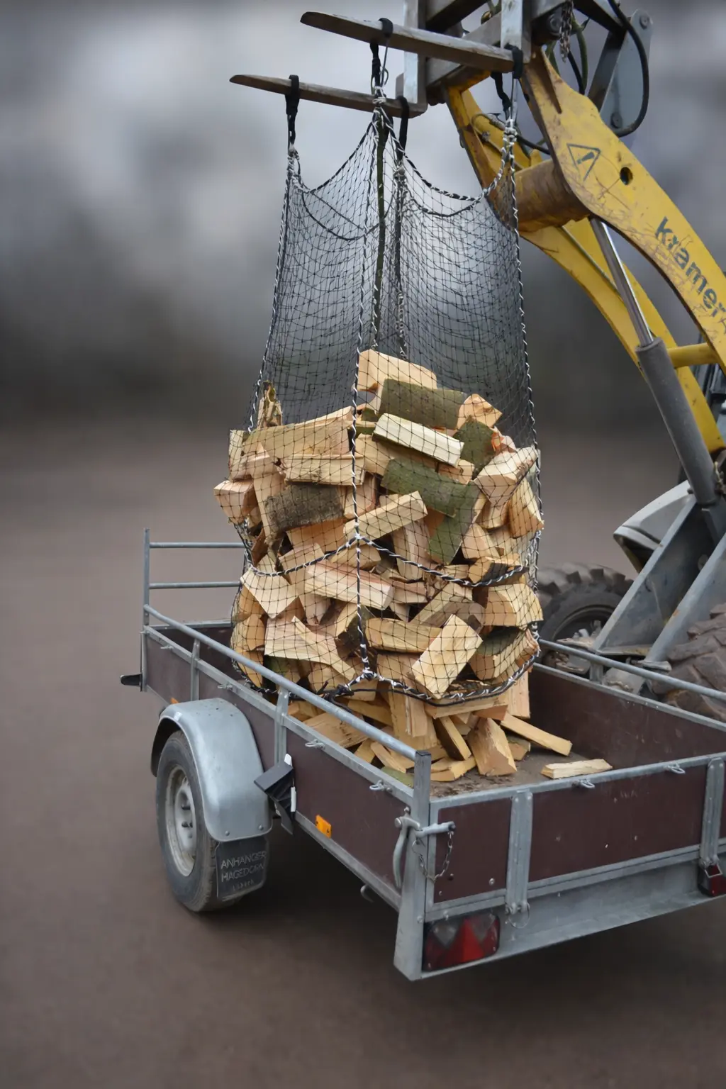 Holzsack mit offenem Boden, gefüllt mit frisch gespaltenem Brennholz, wird per Frontlader in einen Transportanhänger entleert.