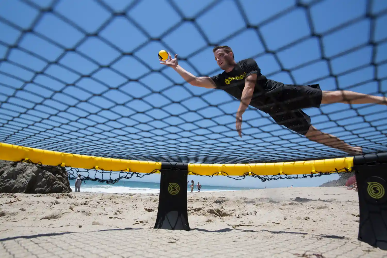 Spikeball‑Spieler springt am Strand, gelb‑schwarzer Ball über dem Netz, dynamische Perspektive