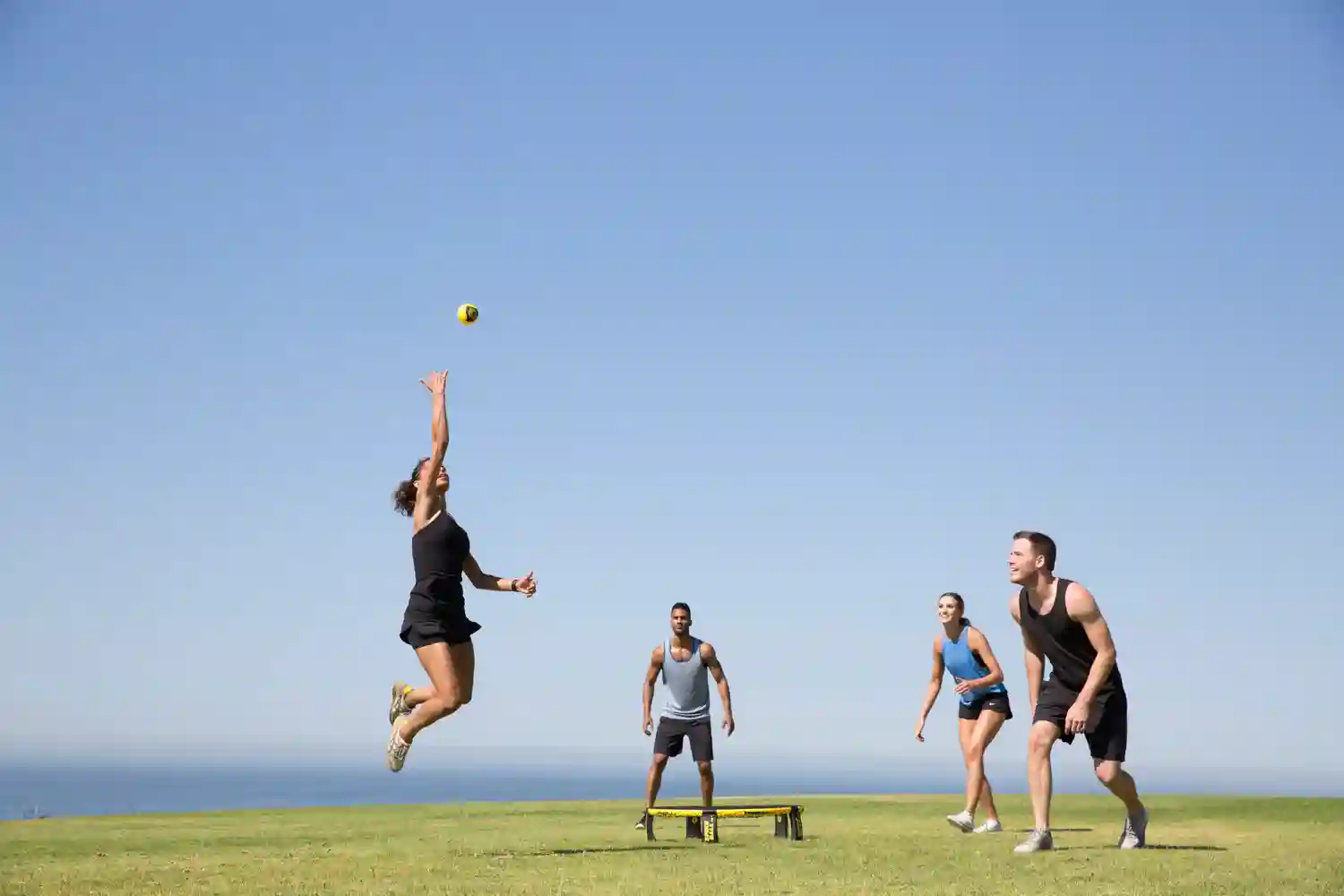 Vier Personen spielen Spikeball auf einer Wiese mit Meerblick, Spielerin springt nach dem Ball