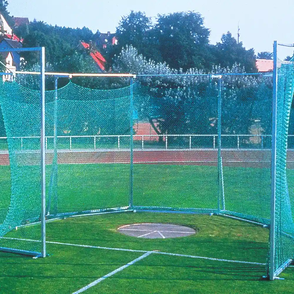 Außenansicht eines Hammerwurfcages auf einem Sportfeld. Das Netz ist an Metallpfosten befestigt, im Inneren liegt ein  betonierter Wurfring. Im Hintergrund befinden sich eine Rasenfläche, eine Laufbahn, Bäume und Gebäude.
