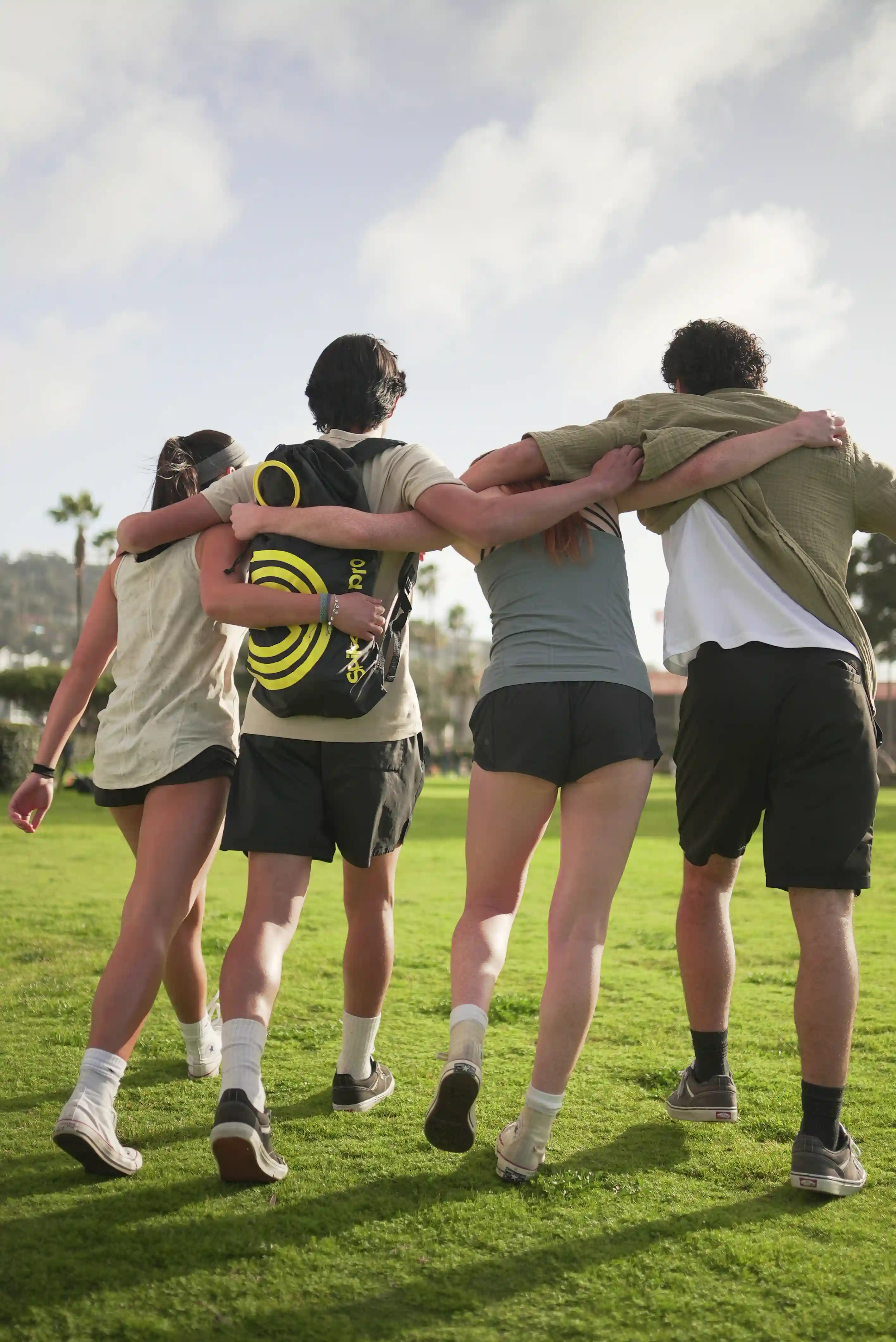 Vier Freunde gehen Arm in Arm über eine Wiese, Rucksack mit Spikeball‑Logo sichtbar