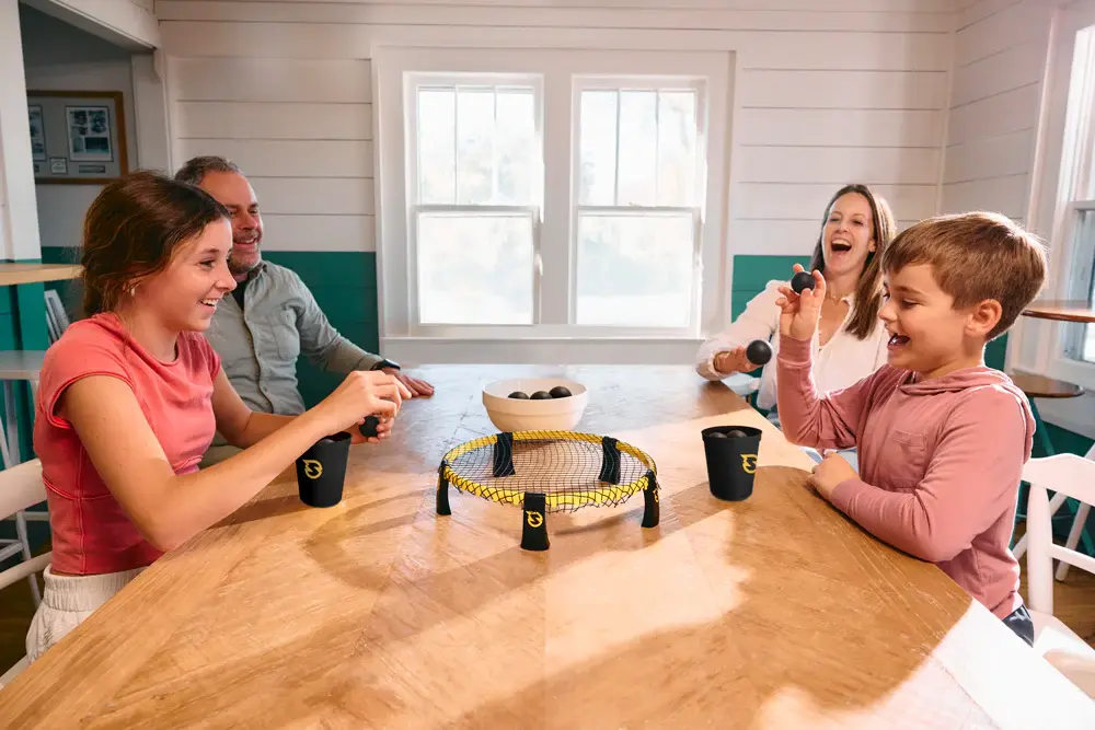 Familie spielt Spikeball Tischspiel an einem Holztisch in heller Wohnung, lachend und aktiv – schwarzes Netz mit gelber Umrandung, schwarze Becher und Bälle, Indoor‑Freizeitspiel für Kinder und Erwachsene